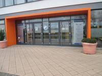 Breite Glasfront mit zwei zweiflügeligen Eingangstüren, umgeben von einem orangefarbenem Rahmen, der ein Vordach bildet. Links und rechts vom Eingang große Pflanzenkübel auf einer gepflasterter Fläche 