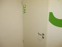 weiße einflügelige Tür in einer weißen Wand, auf der Tür ist ein grüner Schriftzug WC