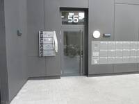Eine Glastür mit einem dunklem Rahmen in einer dunklem Wand. Über der Tür steht in großen, weißen Ziffern auf dem Oberlicht die Nummer 56. Links von der Tür sind 4 Schilder im Querformat übereinander angebracht, die anzeigen, in welchem Stockwerk welche Einrichtung oder Praxis liegt.