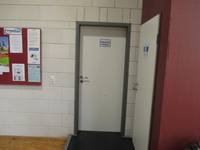 einflügelige Tür mit dunklem Rahmen in einer weißen Wand, links Memotafel mit Aushängen, rechts eine weitere Tür