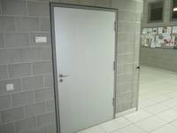 hellgraue Tür in einer grauen Wand, links von der Tür ein kleines Schild. Flurbereich ist hell gefliest