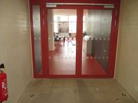 zweiflügelige Glastür mit einem roten Rahmen, vor dem Eingang Fliesen, innen roter Boden. Links vor der Tür hängt ein Feuerlöscher