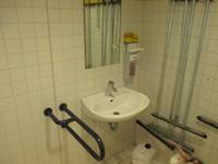 ein Waschbecken mit Spiegel in einer gekacheltten Wand, rechts davon ein Haltegriff, links ein \\\\\\\'Seifenspender und Putzgerät