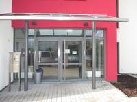 Glasfront mit zweiflügligen Tür, Glasvordach über Eingangsbereich. Die Wand um den Eingangsbereich ist rot gestrichen