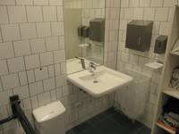  weißes Waschbecken an einer weiß gekachelten Wand, darüber ein Spiegel. Rechts an der Wand ein Handtuchspender, Seifenspender und Mülleimer. Links ein weiterer Mülleimer