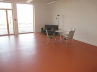 Raum mit einem orangeroten Boden, hinten in der Wand ist eine Glasfront. In der rechten Ecke steht ein Sofa mit einer Stehlampe. Etwas weiter vorne ist ein kleiner Tisch mit drei Stühlen