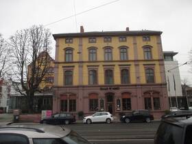 Ein mehrstöckiges gelbliches Gebäude direkt an einer Straße. In der Mitte ist ein Eingang mit Stufen davor.