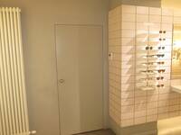 Eine helle Tür in einer hellen Wand, links daneben hängt ein hoher Heizkörper. Links von der Tür hängen 8 kleine Ablagebretter mit Brillengestellen darauf