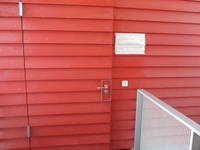 rot lackierte Wand mit waagrechten Brettern, Tür darin eingelassen, rechts von Tür Schild