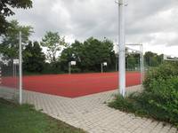 Roter Tartanboden, umgeben von einem gepflasterten Weg. Auf dem Platz sind mehrere Basketballkörbe. Im Hintergrund stehen Bäume