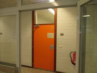 orangefarbene Tür in einer weißen Wand, über der Tür ist eine Scheibe, rechts davon ist an der Wand ein Feuerlöscher