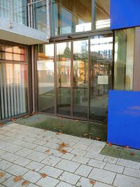 in der Bildmitte: automatische Schiebetür: Auf der Tür stehen die Öffnungszeiten, rechts von der Tür befindet sich ein blauer Würfel mit Fenster, darin befindet sich ein Concierge-Dienst. 