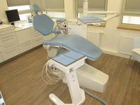 Verstellbarer Behandlungsstuhl , davor verschiebbares Gerät mit Zahnarztinstrumenten. Im Hintergrund niedrige Praxisschränke und PC