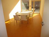 Raum mit  orangefarbenen Boden, Rückseite Glasfront. Im Raum stehen mehrere Tische zu einer Gruppe zusammengestellt, drum herum stehen Stühle