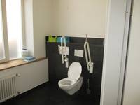 weiße Toilette mit Handlauf rechts und links an einer halbhoch schwarz gefliesten Wand. An der Rückseite ist ein Spültaster. Links davon eine weiße Wand mit einem Fenster