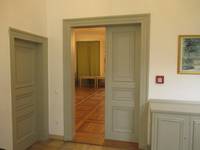 Eine hellgraue Tür mit einem offenstehenden Flügel. Rechts von der Tür steht eine niedrige hellgraue Kommode, darüber hängt ein Bild.
