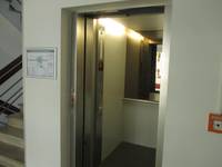 offenstehende Metalltür, dahinter ist die Aufzugskabine mit einem Spiegel an der Rückseite. Rechts ein Feuermelder. Links ein Rettungsplan an der Wand und ein Teil des Treppenhauses.