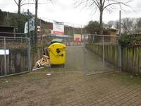 Ein Weg mit einer Steigung führt durch ein Tor in Richtung eines eingezäunten Sportplatz. Links am Weg steht eine gelbe, große Mülltonne.