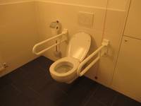 weißes Hänge-WC an  weiß gekachelten Wand. Auf beiden Seiten ist ein Haltegriff