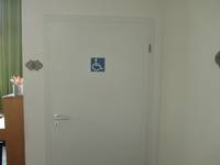 weiße Tür in einer weißen Wand, links neben der Tür und auf der Tür ist ein Rollstuhlsymbol. Links von der Tür ist ein Stück des Raums