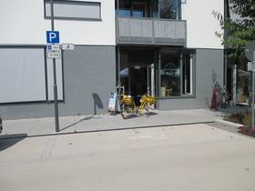 Parkplatz in Längsstellung zur FAhrbahn, im Hintergrund Gebäudeeingang. am Parkplatz Schild mit Rollstuhlfahrer-Symbol