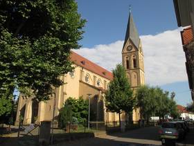Kirchengebäude mit Schrägdach und daran anschließend Turm mit Uhr und Spitzdach, davor eine Wiese mit Bäumen