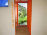 offener Durchgang, links am Durchgang ist vertikal die Anschrift: Ein- und Auszahlungen" in großen Buchstaben angebracht. Die Wand im Raum ist in einem kräftigen Orange gestrichen, an der dem Durchgang gegenüberliegenden Wand ist ein großes, hochformatiges Foto der Neckarwiese mit Blick auf den Heiligenberg aufgehängt.