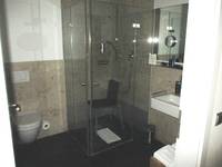 Links im Raum ein Hänge-WC, rechts daneben eine ebenerdige gläserne Duschkabine mit einem Stuhl und einer höhenverstellbaren Handbrause, rechts von der Dusche ist ein Waschtisch mit Spiegel