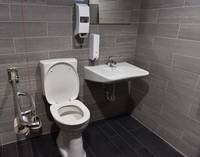 weiße Toilette, links daneben Metallhaltegriff mit WC Papier, rechts daneben weißes Waschbecken. Der Raum ist deckenhoch gefliest. Bodenflisen dunkelgrau, Wandfliesen hellgrau
