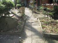 Weg aus quadratischen Betonplatten, jeweils 2 nebeneinander, rechts und links davon sind bepflanzte Flächen oder ein Zaun oder Stufen